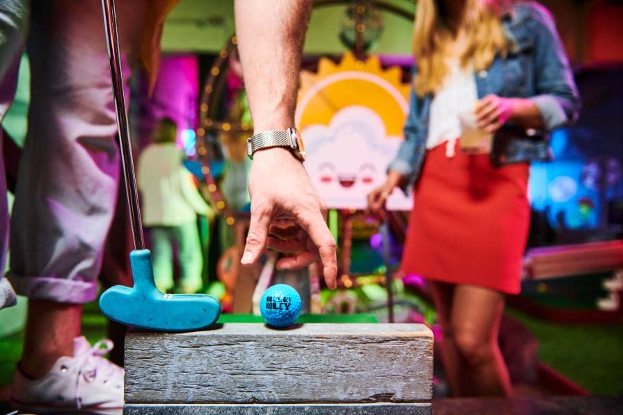 A mini golf ball and putter in teal blue color with the Holey Moley lettering logo in black slapped on the golf ball, with two players and a mini golf course in the background