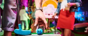 A mini golf ball and putter in teal blue color with the Holey Moley lettering logo in black slapped on the golf ball, with two players and a mini golf course in the background