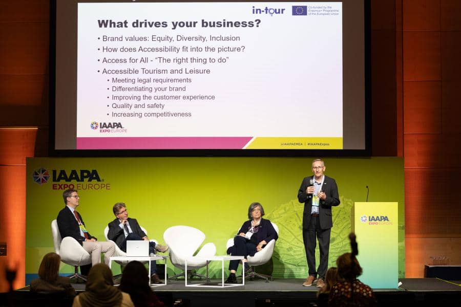 Panelists on stage with a PowerPoint slideshow on a projector screen showing questions on accessible tourism during IAAPA Expo Europe