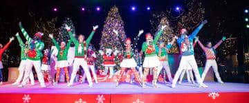 Performer on stage wearing festive holiday costumes and in the center background, Snoopy mascot in a Santa Claus costume also performing at a holiday show at King's Dominion